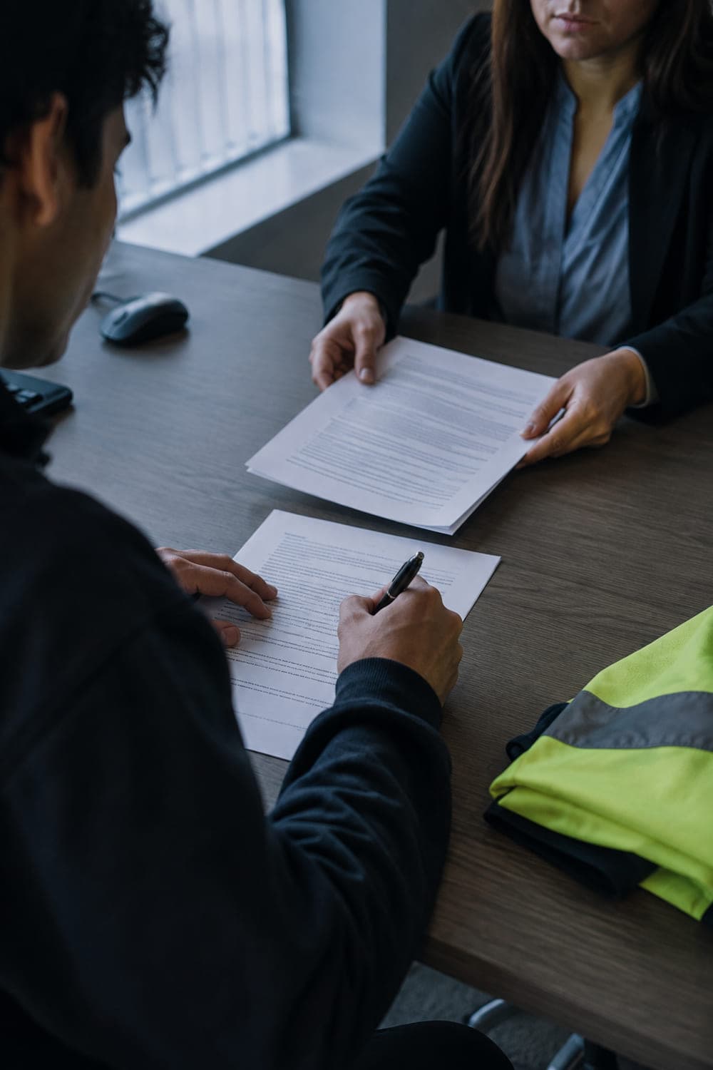 Trabajador firmando su contrato de trabajo frente a la coordinadora de personas, con el chaleco reflectante doblado sobre la mesa