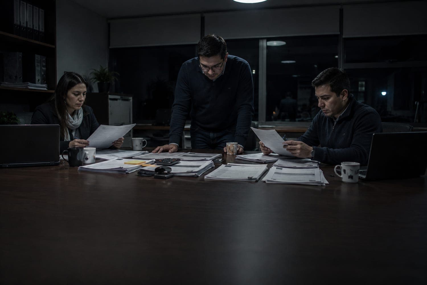 Tres personas armando de noche la carpeta de una licitación pública sobre la mesa de reuniones, con los formularios separados en pilas