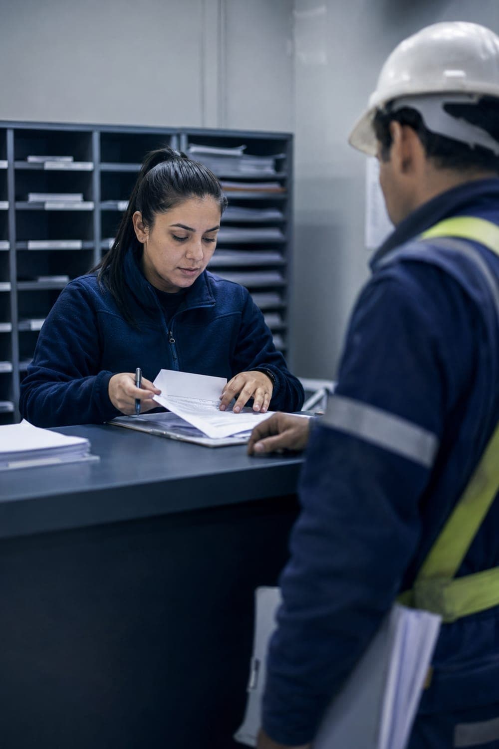 Mostrador de acreditación de contratistas: la encargada revisa documentos con un lápiz en la mano mientras un trabajador con casco espera al otro lado con más papeles