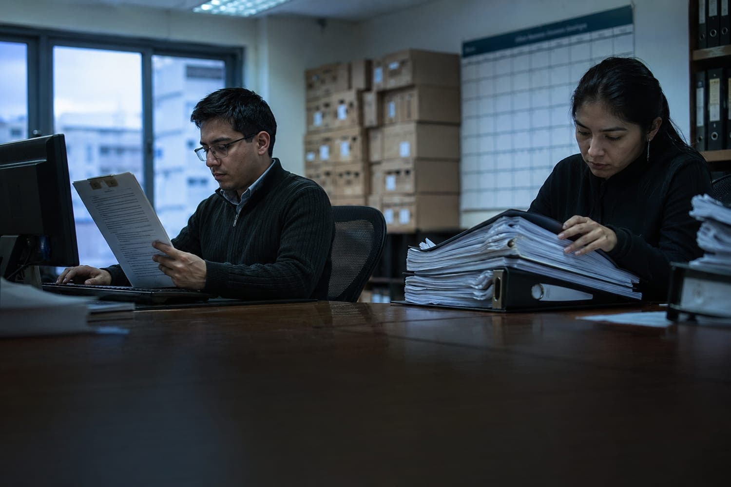 Estudio contable a última hora de la tarde: un contador digita un contrato impreso en el computador mientras su colega revisa un archivador de varios centímetros de grosor
